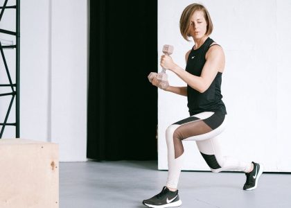 A young woman performing lunges with dumbbells in a modern indoor gym setting.